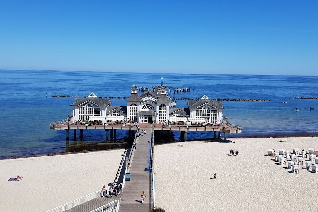 Een steiger die in de zee loopt, met een gebouw aan het einde, een zandstrand en een heldere lucht.