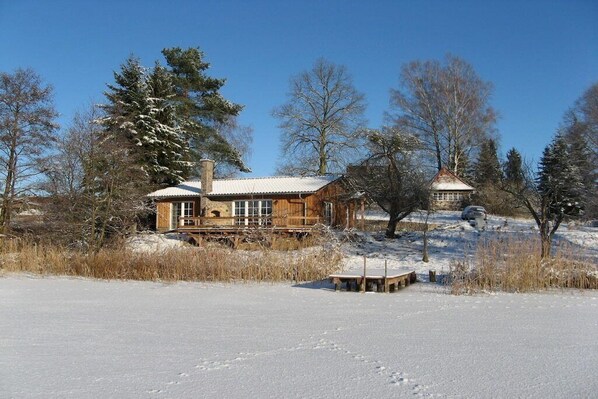 A cabin with a wooden exterior, a chimney, and a small adjacent structure, surrounded by snow-covered ground and trees. - Lakeside idyll Comthurey - Reed House (Wokuhl-Dabelow OT Comthurey)