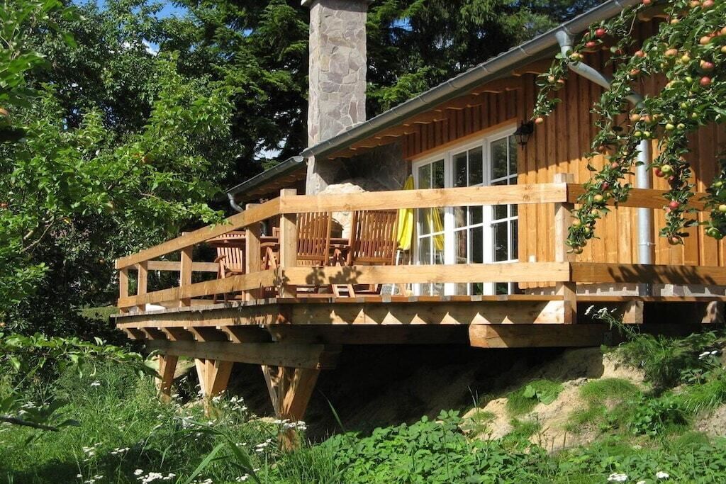 A wooden cabin with a deck, a stone chimney, and a window overlooking greenery.