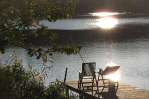 A wooden dock with a chair and a table, overlooking a calm body of water with sunlight reflecting on the surface. - Lakeside idyll Comthurey - Reed House (Wokuhl-Dabelow OT Comthurey)