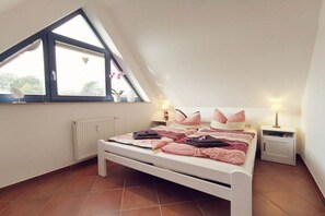 Een slaapkamer met een schuine kap, een bed met roze en witte beddengoed, twee nachtkastjes met lampen en een raam met uitzicht op bomen.