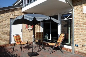 Eine Terrasse mit zwei Stühlen und einem Tisch unter einem großen Sonnenschirm, eine Ziegelwand und eine Glastür, die sich öffnen lässt.