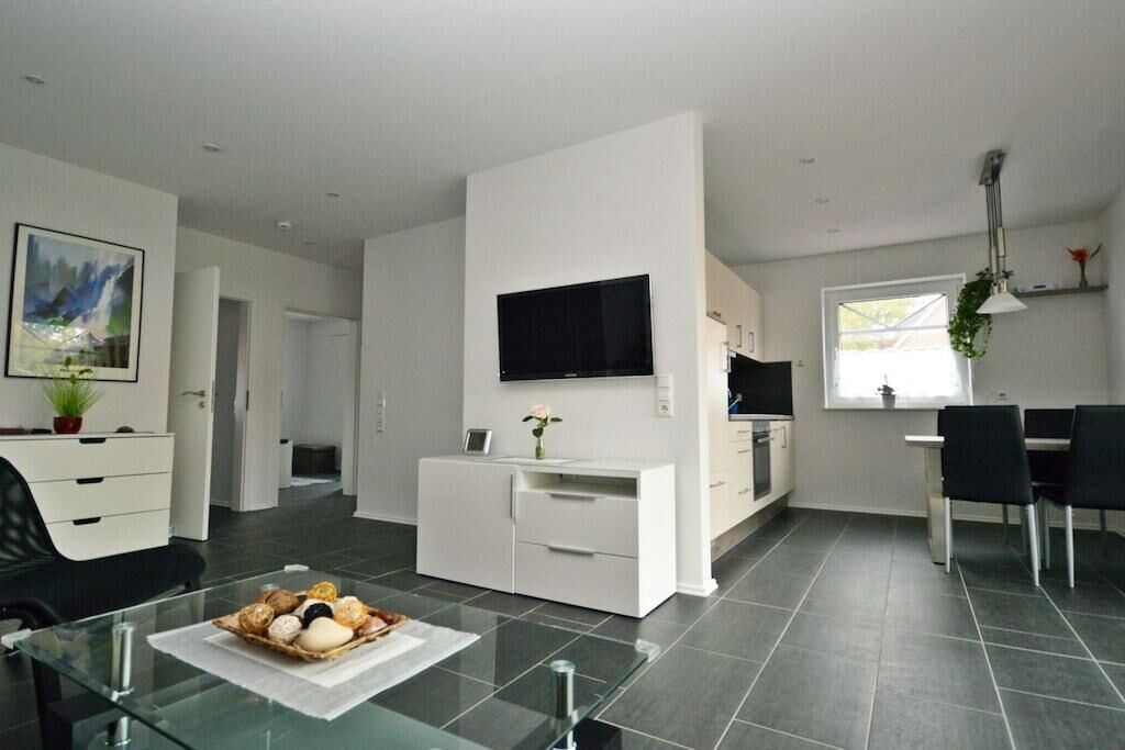 A modern living room with a glass coffee table, a flat-screen TV mounted on the wall, and a dining area with a white table and black chairs.