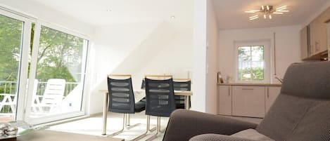 A modern living room with a sofa, dining table, and chairs, a kitchen area with cabinets, and large windows offering a view of the outdoors.