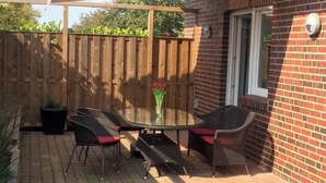A patio with a glass table and chairs, a wooden fence, and a brick house. - Apartment 'Anna Sandbank' with Private Terrace, Private Garden and Wi-Fi (Krummhörn)
