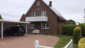 A two-story house with a covered carport, a white fence, and a well-maintained garden. - Apartment 'Anna Sandbank' with Private Terrace, Private Garden and Wi-Fi (Krummhörn)