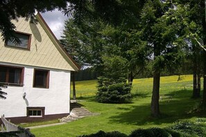 A white building with a sloped roof, a small porch, and a grassy yard with trees and a bench. - Holiday home with 1 bedroom (Eisenbach-Schollach)