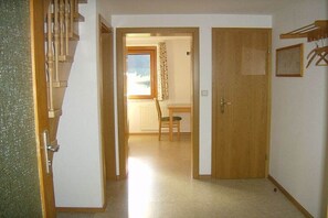 Een hal met een houten trap, een deur en een zicht op een kamer met een stoel en een raam.