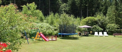A grassy outdoor area with a playground including a slide and swings, surrounded by trees.