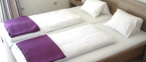 A double bed with white linens and purple accents, a wooden headboard, and a small bedside table with a purple object.