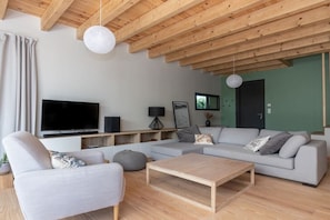 A modern living room with a flat-screen TV, a grey sofa, a wooden coffee table, and a ceiling with exposed beams. - Bioclimat wooden house in the countryside (Messanges)