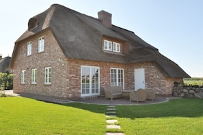 Une maison en briques avec un toit à forte pente, de grandes fenêtres et une terrasse aménagée avec du mobilier d’extérieur.