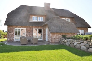Une maison en briques avec un toit de chaume, un mur en pierre et une terrasse aménagée avec du mobilier d’extérieur.