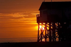 Une structure en bois dotée d’un balcon, qui pourrait être une jetée ou un poste d’observation, avec le soleil couchant en arrière-plan.