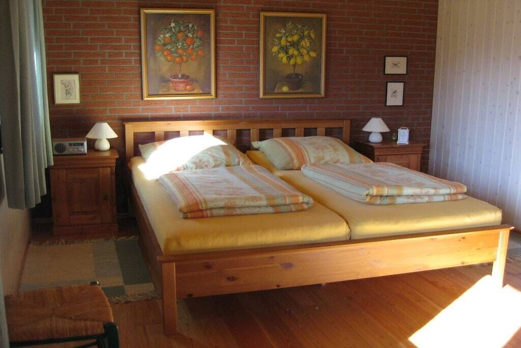 Uma cama de casal com estrutura de madeira e duas almofadas, uma mesa de cabeceira com abajur, e uma parede com duas pinturas emolduradas.