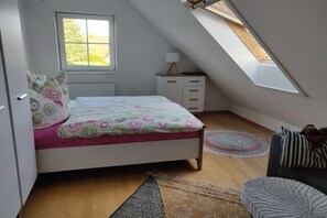 Een slaapkamer met een schuine plafond, een bed met beddengoed met een patroon, een nachtkastje met een lamp, een klein dressoir en een raam met uitzicht op groen.