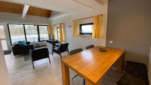 A spacious living area with a wooden dining table, a sofa, and a coffee table. There are large windows with curtains, and a view of the outdoors.