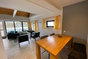 Un vaste espace de vie comprenant une table à manger en bois, un canapé et une table basse. De grandes fenêtres, équipées de rideaux, offrent une vue sur l’extérieur.