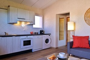 Eine kompakte Küche mit Waschmaschine, Kaffeemaschine und Toaster. Es gibt einen Essbereich mit Tisch, einer Couch und einer Lampe.