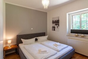 Een hotelkamer met een tweepersoonsbed, een nachtkastje met een lamp, een poster aan de muur en een raam met uitzicht op groen.