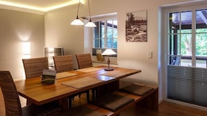 Uma sala de jantar com uma mesa de madeira, cadeiras de fibra natural e um banco. Há uma luminária sobre a mesa, e uma janela com persianas à direita.