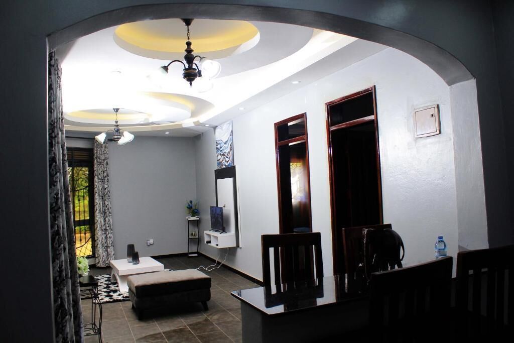 Una sala de estar moderna con un sofá, una mesa de centro y un televisor. Hay un comedor con una mesa y sillas. La habitación tiene un techo con luminarias circulares.