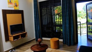 Un salon doté d’un téléviseur à écran plat, d’un meuble de rangement en bois, d’un pouf rond et d’une grande fenêtre ornée de barreaux en fer décoratifs.