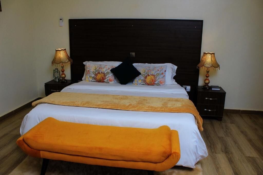 Uma cama com cabeceira escura, duas luminárias laterais e um banquinho no pé da cama.