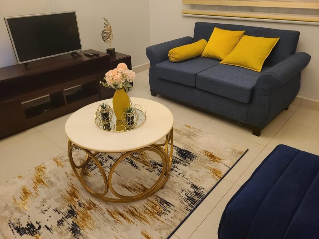 Uma sala de estar com um sofá azul, uma mesa de centro redonda com um vaso e uma televisão em um móvel.