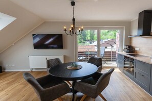 A dining area with a round table and chairs, a chandelier, a television mounted on the wall, and a view of the outside through a glass door. - Small spruce tree with balcony (Braunlage)