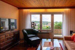 Ein Wohnzimmer mit Fernseher, einer Couch, einem Couchtisch und Blick durch große Fenster.