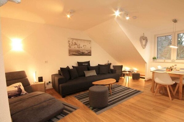 A modern living room with a sofa, ottoman, and a dining area with a table and chairs. - Holiday apartment with 1 bedroom (Braunlage)