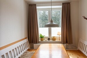 Ein Zimmer mit einem Holztisch, einer Hängelampe, einem Fenster mit Vorhängen und einem Heizkörper unter dem Fenster.