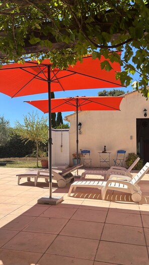 Ein Außenbereich mit einer gefliesten Terrasse, einer Pergola mit roten Sonnenschirmen und Liegestühlen.