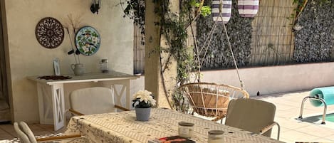 An outdoor patio with a table set for four, surrounded by chairs, under a shaded area.