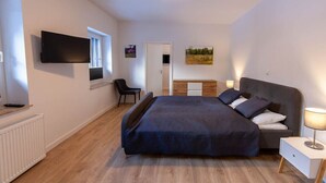 Quarto com cama, mesa de cabeceira, luminária, televisão, cadeira e obra de arte na parede.