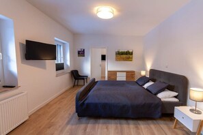 Une chambre à coucher comprenant un lit, une table de chevet, une lampe, un téléviseur, une chaise et une œuvre d’art accrochée au mur.
