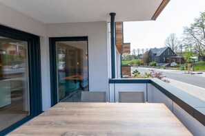 Un balcon moderne avec une table en bois, une porte vitrée donnant sur une pièce et une vue sur une rue résidentielle.