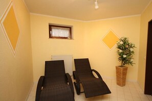 Ein Zimmer mit zwei Liegesesseln, einem kleinen Fenster mit Vorhang, einem Heizkörper und einer Topfpflanze.