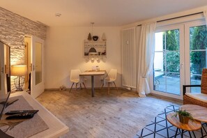 A room with a stone wall, a dining table with chairs, a small shelf with decorative items, and a large window with curtains. - Small mountain suite (Braunlage)