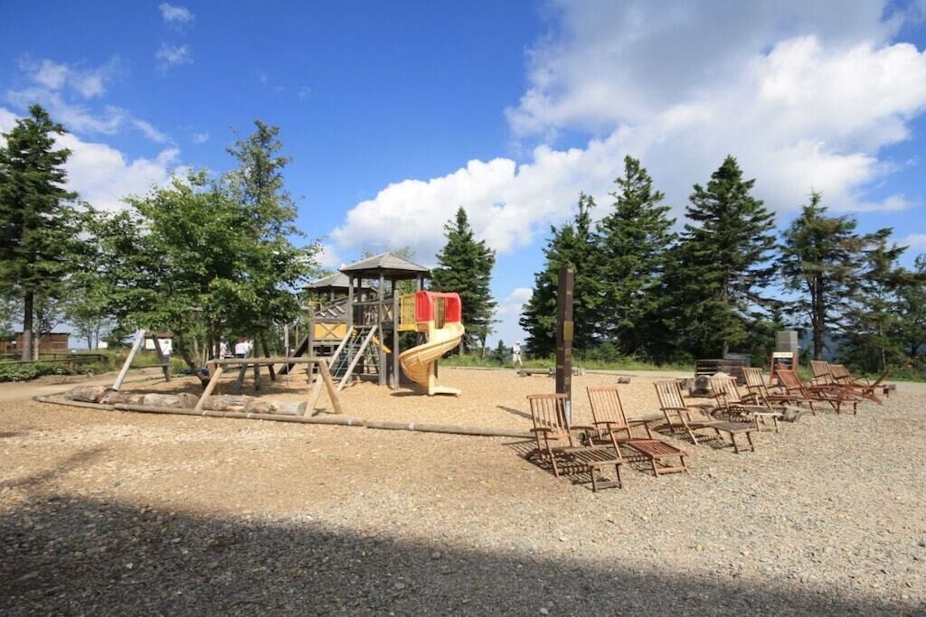 Ein Spielplatz mit Rutsche und Klettergerüsten, umgeben von Bäumen und einem Kiesbereich mit Holzstühlen.