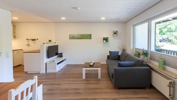 Uma sala de estar moderna com sofá, mesa de centro e TV. É visível uma área de cozinha ao fundo, com armários e uma janela.