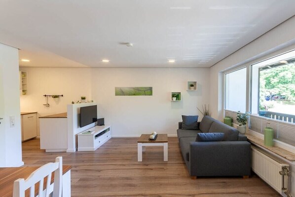 Un salon moderne avec un canapé, une table basse et un téléviseur. On aperçoit, au fond, un coin cuisine avec des placards et une fenêtre.