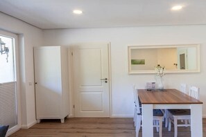 Une pièce avec une table à manger en bois, des chaises blanches, une porte blanche et un miroir au mur.