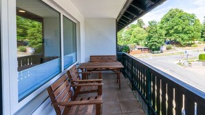 Varanda com bancos de madeira e uma mesa, vista para uma rua com árvores e carros estacionados.