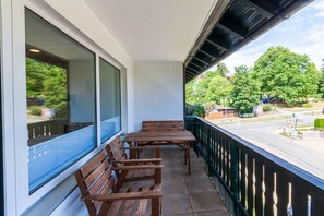 Un balcon avec des bancs en bois et une table, donnant sur une rue bordée d’arbres et où sont garées des voitures.