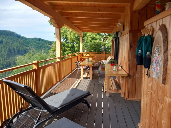 Una terraza de madera con una mesa, sillas y una tumbona, con vistas a un valle boscoso.