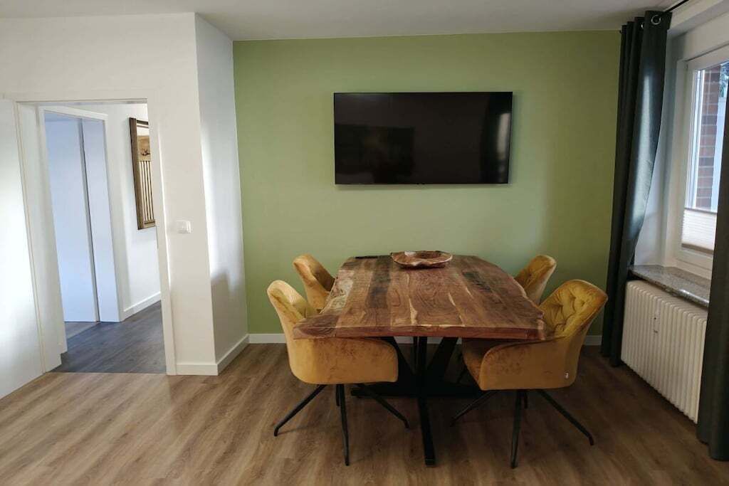 A dining area with a wooden table and four chairs, a flat-screen TV on the wall, and a radiator on the right.