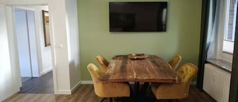 A dining area with a wooden table and four chairs, a flat-screen TV on the wall, and a radiator on the right.