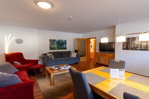 Ein Wohnzimmer mit einer Couch, einem roten Sessel, einem Esstisch mit Tischsets, einem Couchtisch und einem Fernseher.